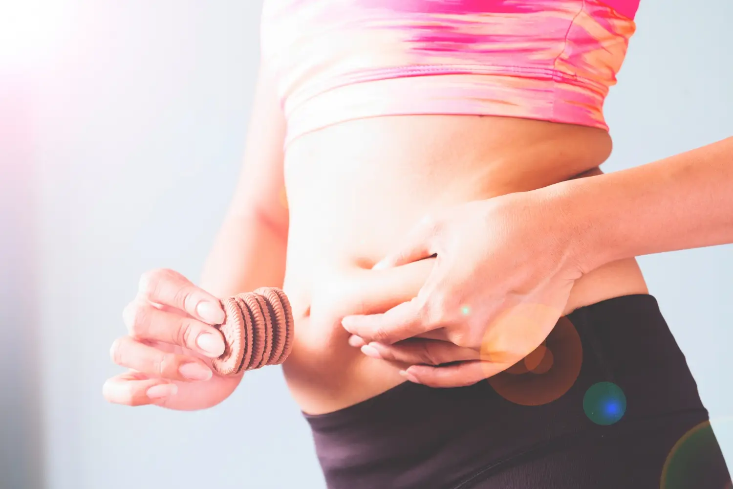 Fitness female in pink sport bra squeeze her fat waist and snack in other hand with copy space